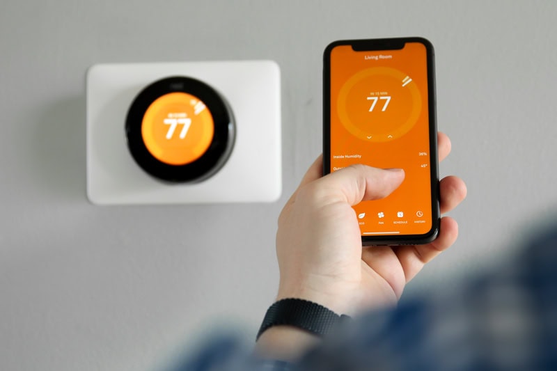 Person adjusting a smart thermostat with a smartphone app to control home temperature and improve energy efficiency.