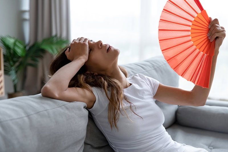 Why Is My AC Blowing Hot Air? Photo of a young girl fanning herself off to cool down on her living room couch.