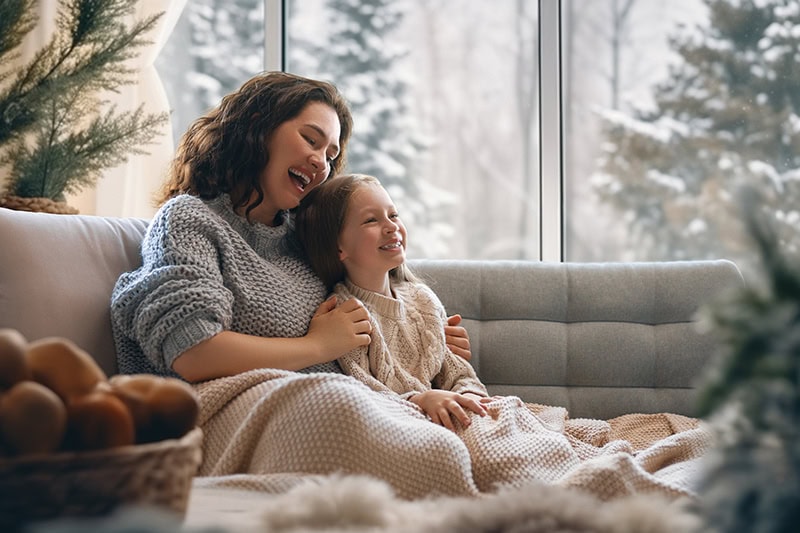 5 Ways to Save on Heating Bills in Portland. Happy loving family. Mother and daughter are hugging and enjoying winter nature in the window.